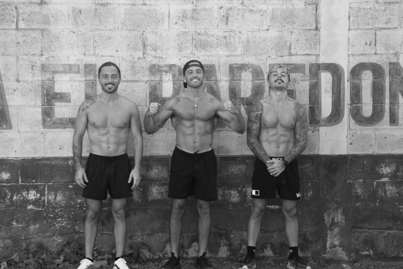 The three brothers standing together against a painted cinderblock wall.