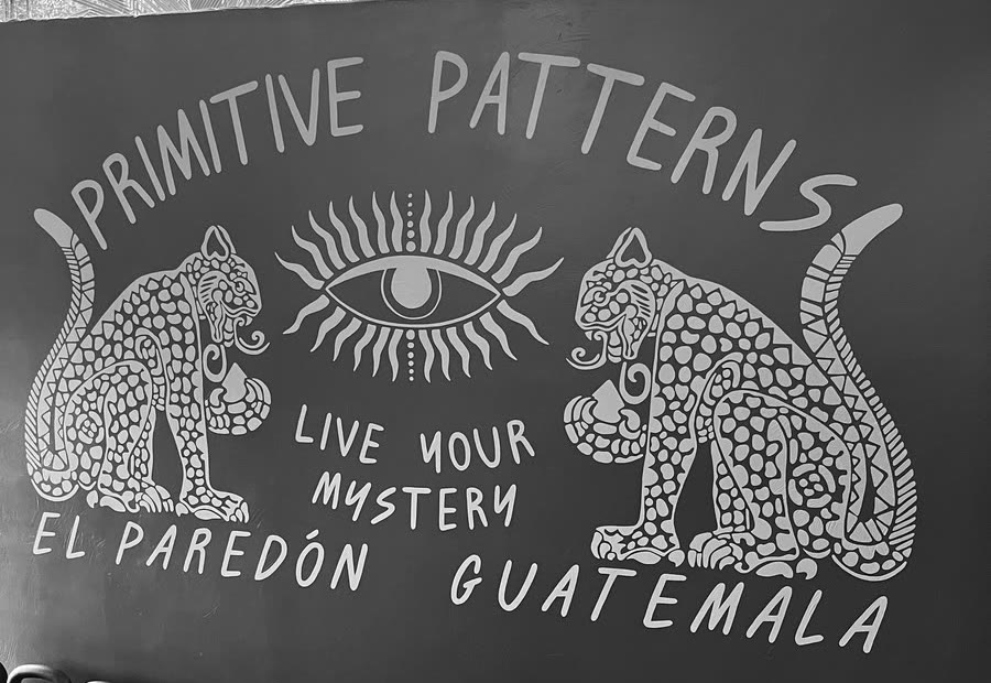 Hand-painted mural reading Primitive Patterns, Live Your Mystery, El Paredón Guatemala, with two jaguars flanking a radiant eye.